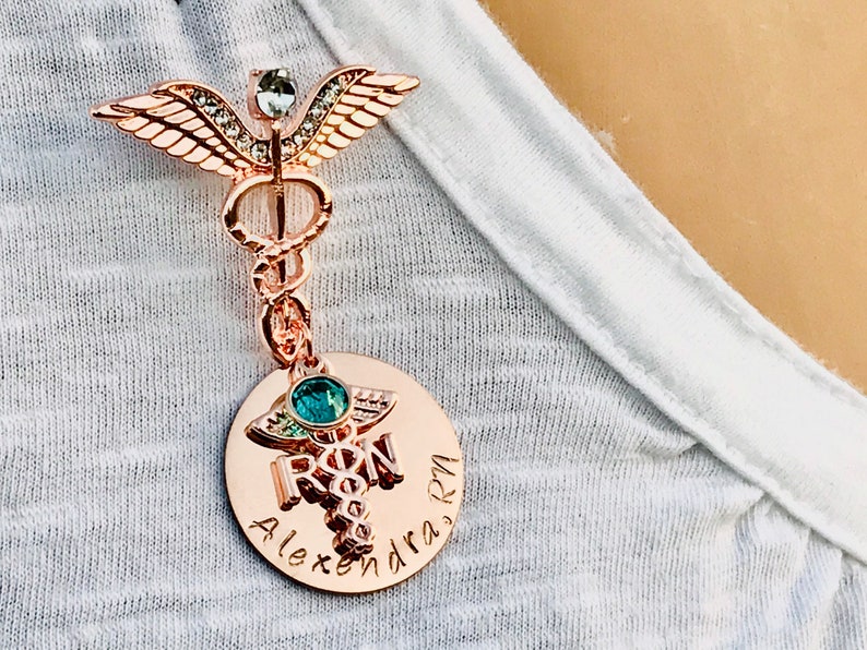 May include: Rose gold medical caduceus pin with a clear crystal and green gem. The pin has a round charm with the words "RN" and "Alexandra" engraved on it.