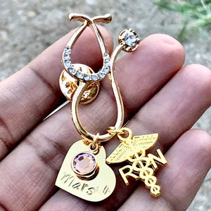 May include: Gold stethoscope pin with a pink crystal heart charm and a gold charm with the letters RN and a caduceus. The heart charm has the name "Marsh" engraved on it.