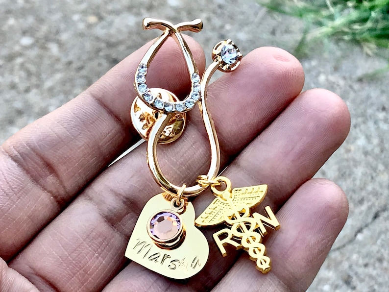 May include: Gold stethoscope pin with a pink crystal heart charm and a gold charm with the letters RN and a caduceus. The heart charm has the name "Marsh" engraved on it.