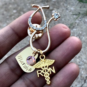 May include: Gold stethoscope charm with rhinestones, a heart charm with the name "Marcia" engraved on it, and a charm with the letters "RN" and a caduceus symbol.