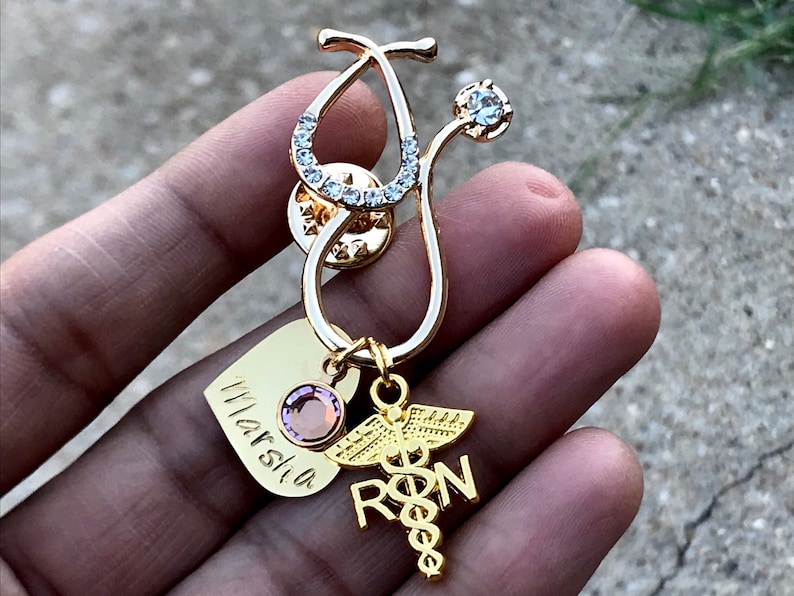 May include: Gold stethoscope charm with rhinestones, a heart charm with the name "Marcia" engraved on it, and a charm with the letters "RN" and a caduceus symbol.