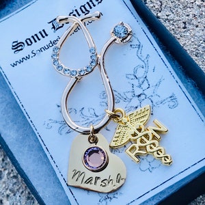 May include: Gold stethoscope charm with a pink heart charm engraved with the name "Marsha" and a gold charm with the letters "RN" and a caduceus symbol. The charms are in a black gift box with a white label that says "Sonu Designs" and "www.Sonudesigns.com".