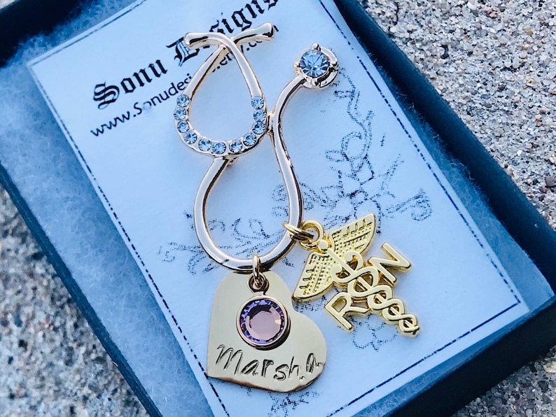May include: Gold stethoscope charm with a pink heart charm engraved with the name "Marsha" and a gold charm with the letters "RN" and a caduceus symbol. The charms are in a black gift box with a white label that says "Sonu Designs" and "www.Sonudesigns.com".