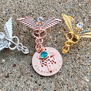 May include: Three gold, silver, and rose gold medical caduceus pins with rhinestones. The rose gold pin has a round charm with the word "RN" and a green gemstone.
