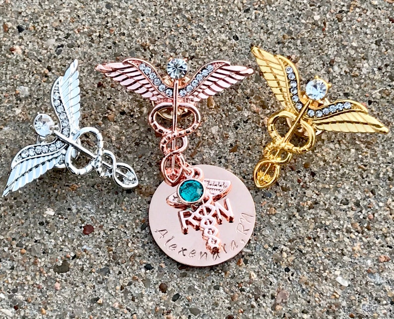 May include: Three gold, silver, and rose gold medical caduceus pins with rhinestones. The rose gold pin has a round charm with the word "RN" and a green gemstone.