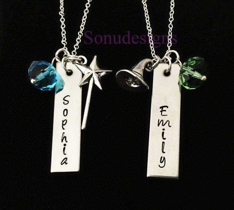 Może przedstawiać: Dwa srebrne naszyjniki z spersonalizowanymi zawieszkami z imionami, jeden z niebieskim kryształem i zawieszką w kształcie gwiazdy, drugi z zielonym kryształem i zawieszką w kształcie kapelusza czarownicy. Zawieszki z imionami "Sophia" i "Emily".