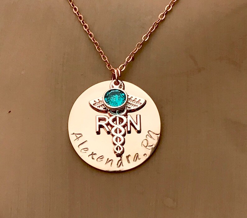 May include: Rose gold necklace with a round pendant featuring a medical caduceus symbol, the letters "RN" and the name "Alexandra RN". A small turquoise gemstone is set above the caduceus.