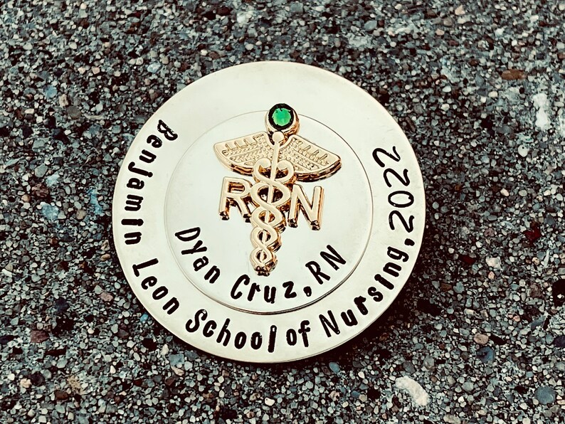 Peut inclure: &Eacute;pinglette ronde dor&eacute;e avec un bijou vert et le texte "Benjamin Leon School of Nursing, 2022" et "Dylan Cruz, RN" avec un symbole de caduc&eacute;e.