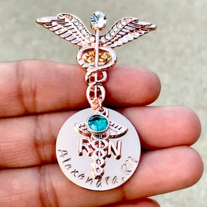 May include: Rose gold medical caduceus pin with a clear crystal and a teal blue stone. The pin has a round charm with the words "RN" and "Alexandra RN" engraved on it.