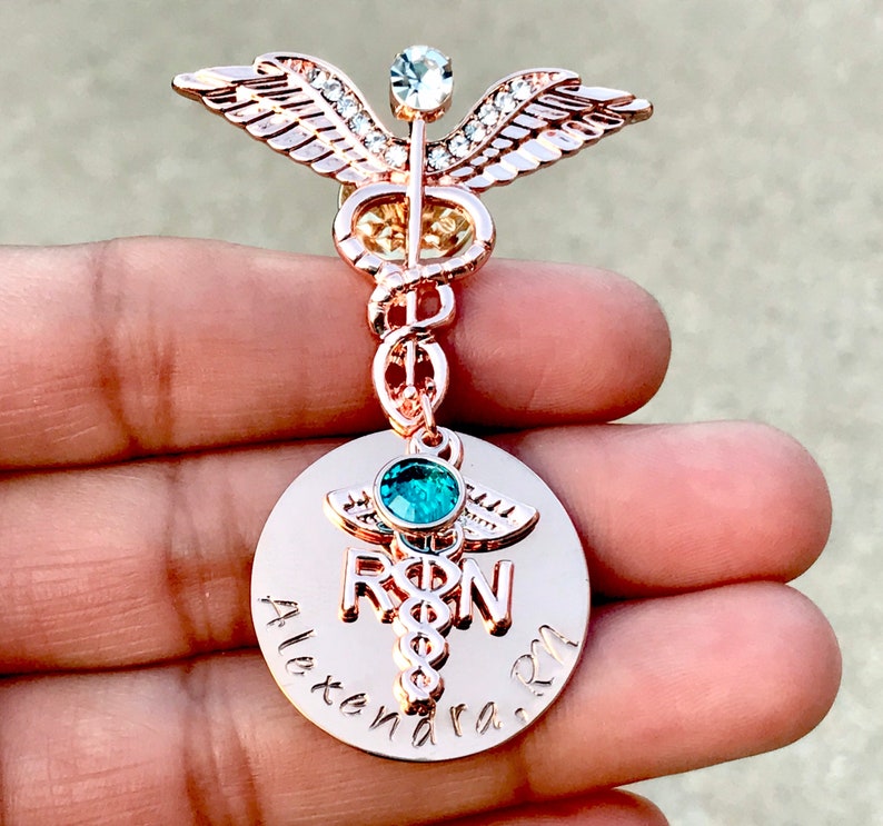 May include: Rose gold medical caduceus pin with a clear crystal and a teal blue stone. The pin has a round charm with the words "RN" and "Alexandra RN" engraved on it.