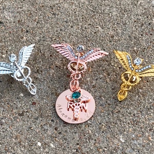 May include: Three gold-toned medical caduceus pins with rhinestones. The center pin has a round charm with a green stone and the text "RN" and "Alexandra 2021".