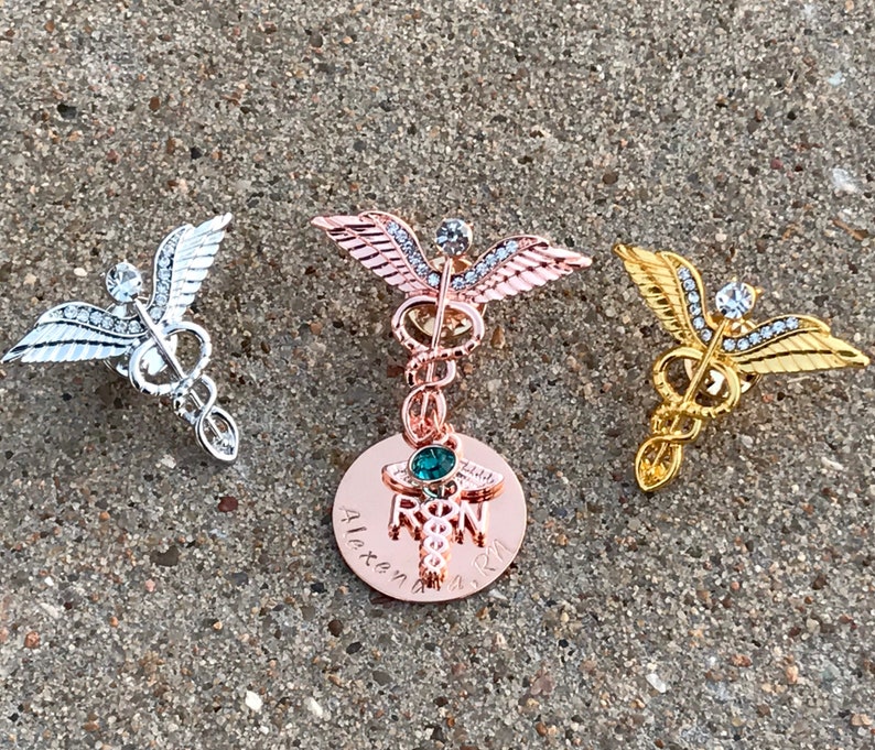 May include: Three gold-toned medical caduceus pins with rhinestones. The center pin has a round charm with a green stone and the text "RN" and "Alexandra 2021".