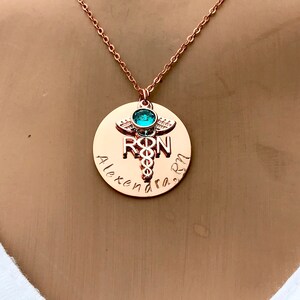 May include: Rose gold necklace with a round pendant featuring a medical caduceus symbol, the letters "RN" and the name "Alexandra" engraved on it. A turquoise gemstone is set at the top of the caduceus.