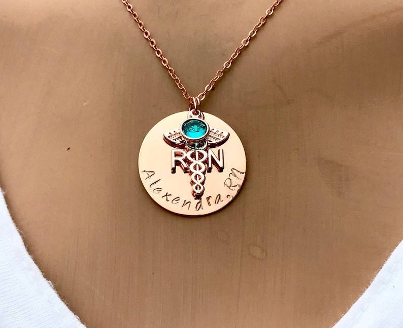 May include: Rose gold necklace with a round pendant featuring a medical caduceus symbol, the letters "RN" and the name "Alexandra" engraved on it. A turquoise gemstone is set at the top of the caduceus.