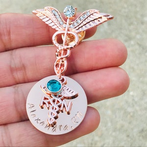 May include: Rose gold medical caduceus pin with a clear crystal and a round charm with a blue crystal and the text "RN" and "Alexandra RN".