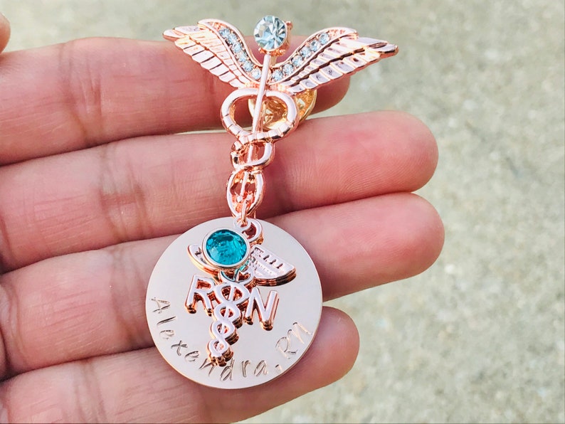 May include: Rose gold medical caduceus pin with a clear crystal and a round charm with a blue crystal and the text "RN" and "Alexandra RN".
