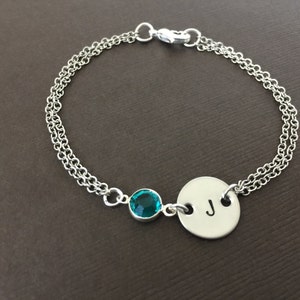 May include: Silver-toned bracelet featuring a teal gemstone and a circular charm with the letter "J". The bracelet has a double chain design and a clasp closure. A personalized jewelry item.