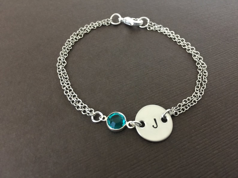 May include: Silver-toned bracelet featuring a teal gemstone and a circular charm with the letter "J". The bracelet has a double chain design and a clasp closure. A personalized jewelry item.