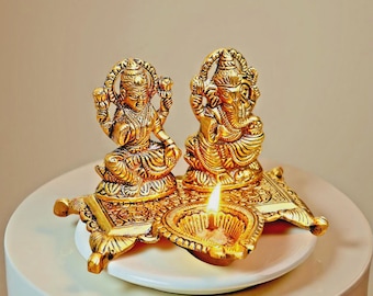 May include: A golden religious display featuring two detailed statues and a lit oil lamp. The statues depict seated figures with ornate headdresses and jewelry. The lamp's flame casts a warm glow on the gold-colored metal.