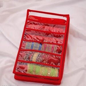 May include: A red rectangular jewelry organizer case featuring multiple horizontal clear plastic compartments. The organizer is filled with various sets of stacked bangles in blue, gold, pink, and green colors, visible through the transparent windows.