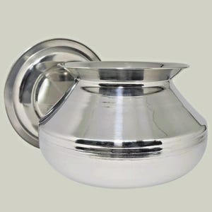 以下が含まれることがあります： 丸みを帯びたボディと幅広のフレアリムを備えた、光沢のある銀色の金属製の鍋。鍋は円形の平らなベースに取り付けられています。表面は光を反射し、滑らかな質感を際立たせています。このキッチン用品はおそらく調理に使用されます。