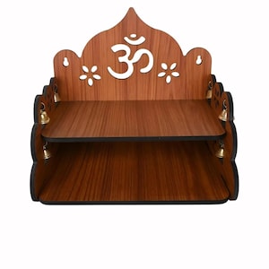 Op de afbeelding: Een houten gebedsplank met een donkerbruine afwerking. De plank heeft twee niveaus en is versierd met een wit Om-symbool, bloemuitsparingen en kleine gouden belletjes. Het ontwerp is symmetrisch en geschikt voor religieuze items.