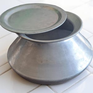 May include: A silver cooking pot with a lid, slightly open. The pot has a rounded base and a wide opening, suggesting it's for cooking. The lid rests on the rim, showing the inside. The pot is on a white tiled surface, typical of a kitchen.