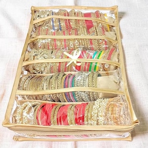 May include: A rectangular, gold-trimmed jewellery storage case with a clear plastic top, featuring four internal padded rods organised to hold numerous stacked bangles in various colours including red, gold, green, and blue with intricate metallic detailing.
