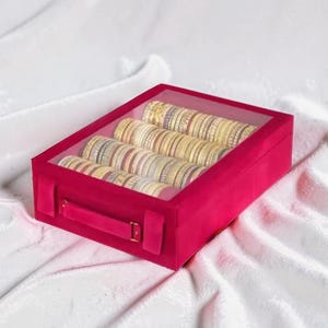 May include: A rectangular, hot pink velvet storage box with a clear, transparent lid. Inside, the box contains three rows of stacked, metallic gold and silver bangles, each featuring intricate, thin, textured patterns along their surfaces.