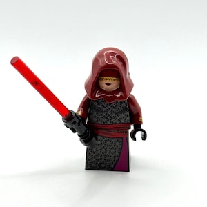 Può includere: Una minifigure LEGO che indossa un cappuccio rosso scuro e una veste nera con un motivo geometrico grigio e una fascia rossa. Il personaggio ha una benda dorata sugli occhi e tiene in mano una spada laser in plastica rossa traslucida.