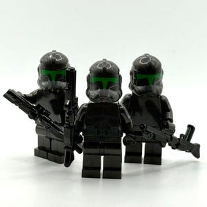 Peut inclure: Trois figurines de soldats en plastique noir avec des casques noirs assortis dotés de visières vert vif. Chaque figurine est équipée d'un accessoire de fusil blaster en plastique noir tenu dans les mains, debout en groupe.