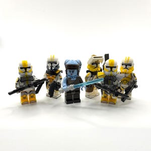 Può includere: Sei minifigure Lego. Cinque indossano armature bianche e gialle con fucili neri. Una ha la pelle e i capelli blu, con una spada laser blu. Su sfondo bianco.