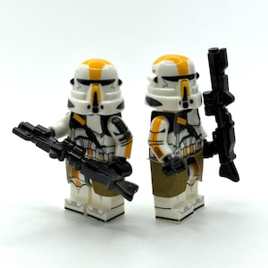 May include: Two plastic toy soldier minifigures featuring white armour with yellow accents and black detailing. Each figure wears a tan fabric kama around the waist and holds a black plastic blaster rifle. The helmets have a distinct yellow stripe.