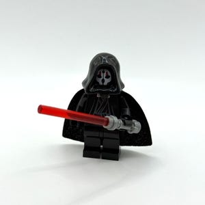 May include: A black plastic minifigure wearing a hooded cloak and a mask with white and red facial details. The figure holds a silver-handled lightsaber with a translucent red blade, featuring a black torso with printed grey lines.