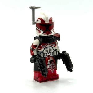 Könnte beinhalten: Eine Spielzeug-Soldaten-Minifigur mit einem weiß-roten Helm mit grauer Antenne, einem schwarzen Halsteil sowie weiß-roter Rüstung. Die Figur hält zwei schwarze Kunststoff-Blaster in den Händen und trägt einen rot-schwarzen Stoffumhang.