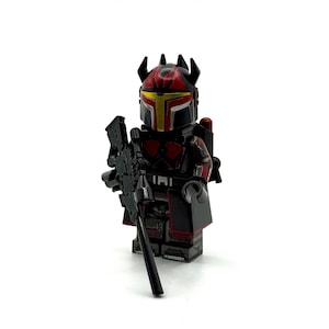 Könnte beinhalten: Eine schwarz-rote Kunststoff-Minifigur mit einem Helm mit spitzer Krone und gelbem Visier. Die Figur verfügt über detaillierte Rüstungsaufdrucke, einen schwarzen Umhang und hält ein langes, schwarzes Scharfschützengewehr in der Hand.