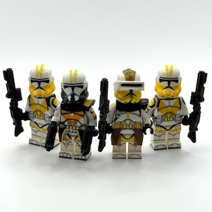 May include: Four plastic toy soldier minifigures with white and yellow armour. Each figure features a unique helmet design, including one with a visor. They are equipped with black plastic blasters held in their hands. The figures stand in a row.