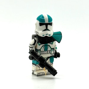 Pode incluir: Uma minifigura de soldado de brinquedo em plástico com capacete e armadura brancos com detalhes em verde-azulado. A figura usa uma ombreira de tecido preto e segura um rifle blaster de plástico preto com marcações detalhadas em preto e verde-azulado.