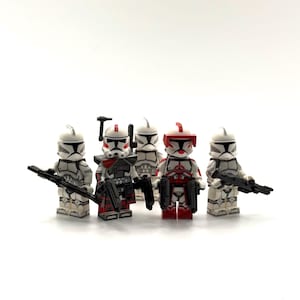 Peut inclure: Cinq figurines blanches et grises avec des détails rouges et noirs, chacune tenant une arme noire. Les figurines sont disposées en rangée sur un fond blanc. Les figurines portent des casques.