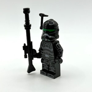 May include: A black plastic minifigure featuring a helmet with a green visor, detailed armour printing, and a long black rifle accessory. The figure includes a small black antenna piece attached to the side of the helmet, standing upright.