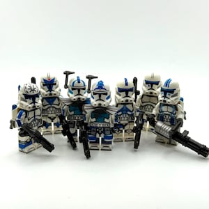 May include: A collection of seven plastic toy soldier minifigures featuring white armor with blue markings and patterns. Each figure wears a distinct helmet design and carries a black plastic weapon, including rifles and a heavy rotating blaster cannon.