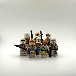 Puede incluir: Un grupo de minifiguras de Lego Star Wars, que incluyen soldados clon con armadura blanca y naranja, y una figura Jedi. Las figuras están de pie juntas, algunas sosteniendo blasters negros, sobre un fondo blanco. Los soldados clon tienen cascos.