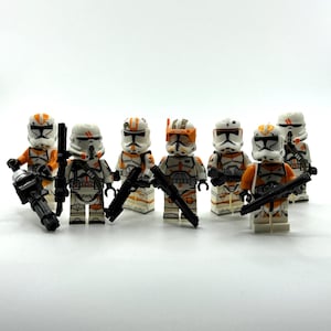 Könnte beinhalten: Eine Sammlung von sieben Spielzeugsoldaten-Minifiguren aus Kunststoff, die jeweils eine weiß-orangefarbene Rüstung mit schwarzen Details tragen. Die Figuren sind mit verschiedenen schwarzen Spielzeug-Blastern und Waffen ausgestattet.