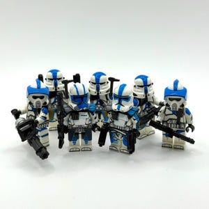 Op de afbeelding: Een verzameling van zeven plastic bouwsteen-minifiguren met wit en blauw pantser en gedetailleerde helmontwerpen. Elk figuur is uitgerust met zwarte plastic speelgoedblasters of zware wapens en staat in een groepsformatie.