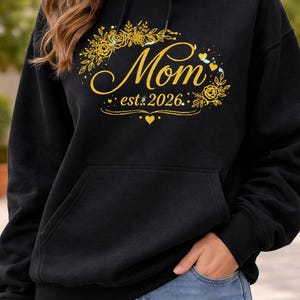 Puede incluir: Sudadera con capucha negra con bolsillo frontal, con un diseño floral dorado y la palabra "Mom" en escritura cursiva. Debajo está el texto "est. 2026". El diseño incluye corazones y pequeños elementos decorativos.