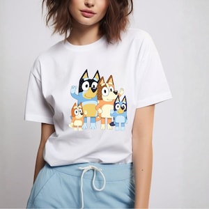 May include: White t-shirt featuring a colorful graphic of a cartoon family of four dogs. The dogs are in shades of blue, orange, and brown. The shirt is a crew neck style.