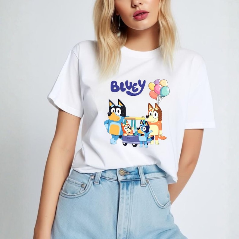 May include: White t-shirt featuring the word "Bluey" above a cartoon image of the Bluey family with balloons. The cartoon characters are in shades of blue, orange, and brown. The shirt is made of a soft material.