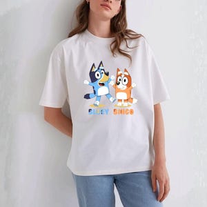 May include: White t-shirt featuring a cartoon illustration of two dogs, one blue and one orange, with the text "Bluey Bingo" in blue and orange. The dogs are in a playful pose with their arms raised. The shirt has short sleeves and a crew neck.
