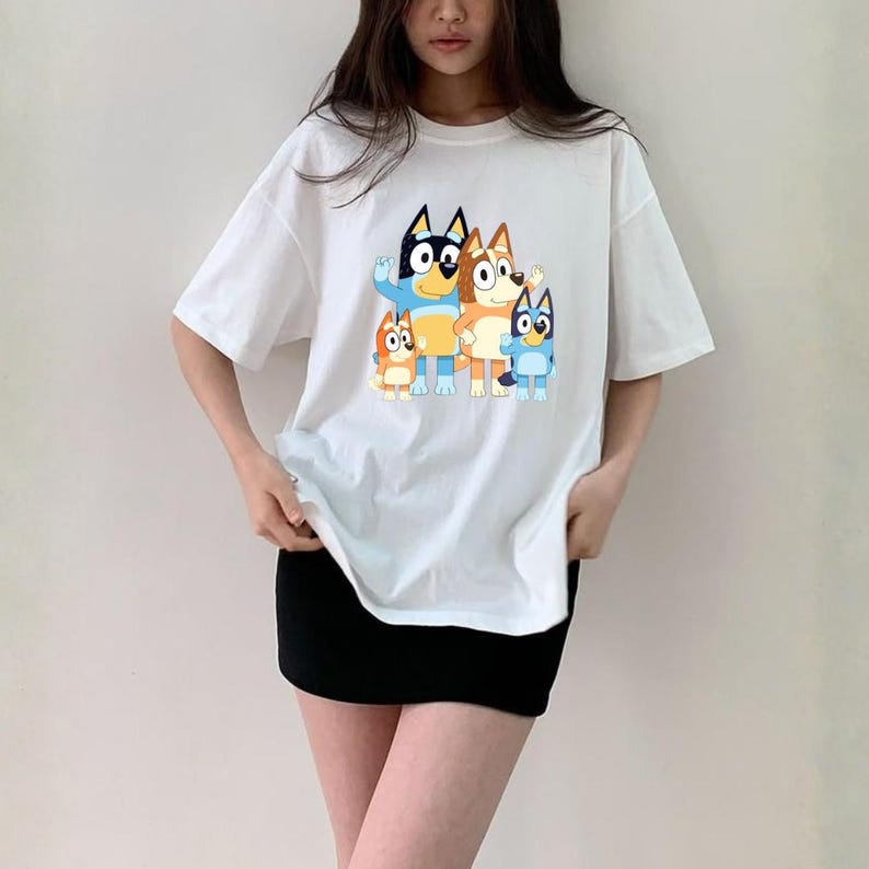 May include: White t-shirt featuring a cartoon family of blue and orange dogs. The graphic is centered on the front of the shirt. The shirt has short sleeves and a crew neck.