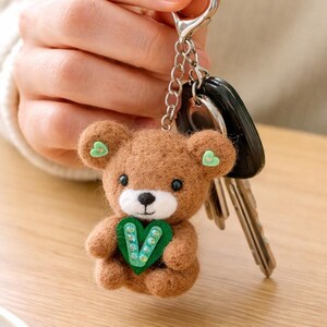 Può includere: Un portachiavi a forma di orsetto in feltro marrone con un accento a forma di cuore verde. L'orso ha accenti verdi a forma di cuore sulle orecchie e una catena d'argento con chiavi attaccate. Il portachiavi è tenuto da una persona.